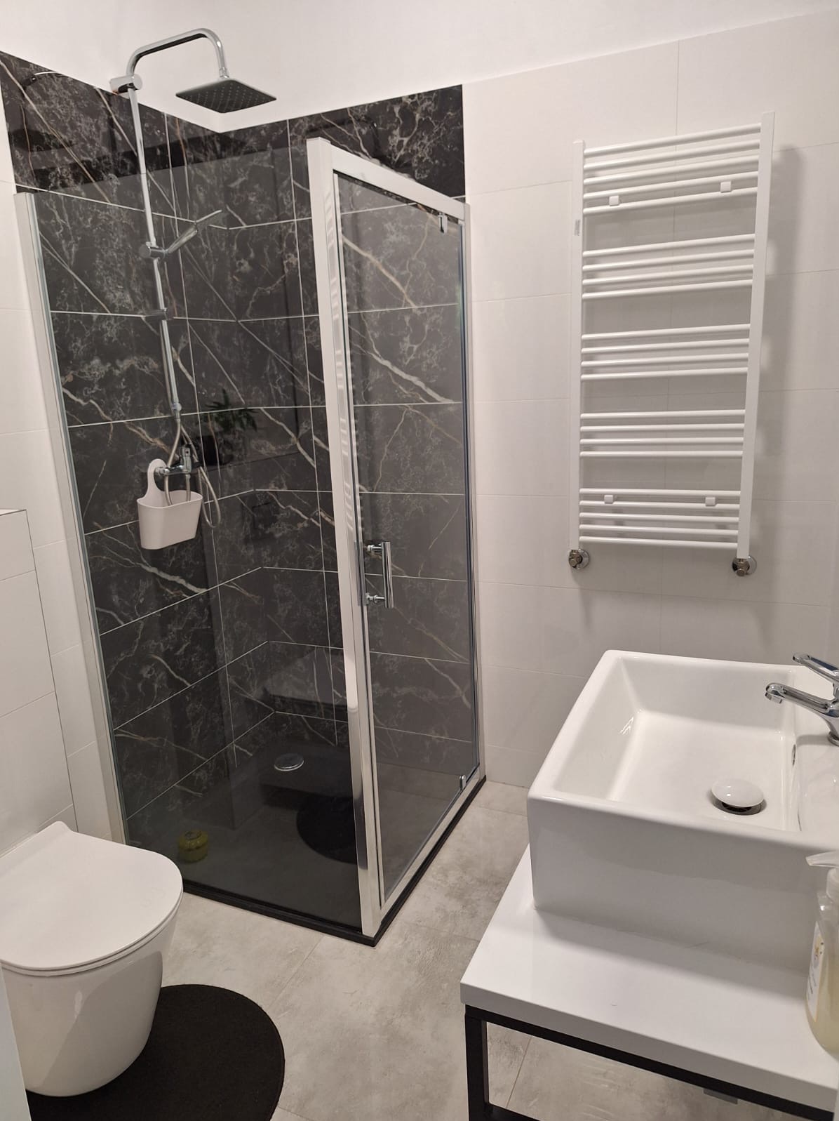 Nowoczesna łazienka z kabiną prysznicową wyłożoną czarnymi płytkami imitującymi marmur, białą umywalką nablatową i grzejnikiem drabinkowym na białej ścianie.