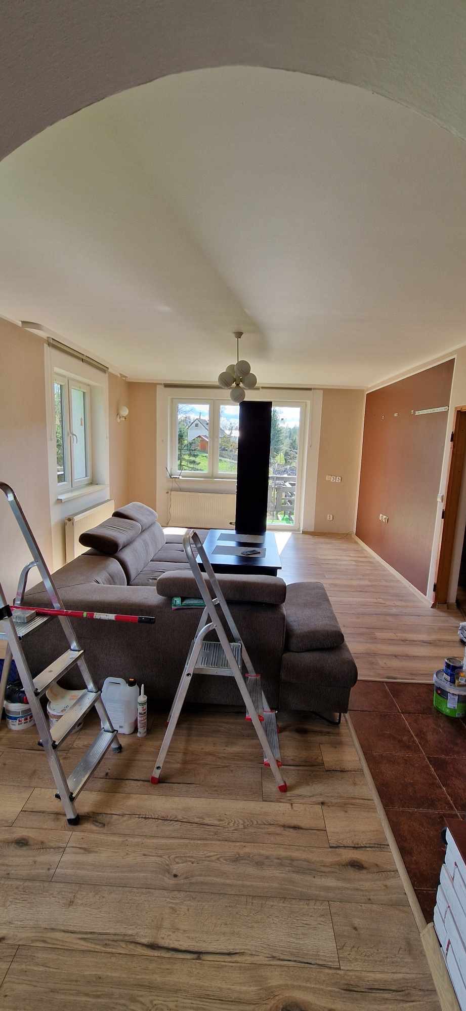 Salon w trakcie malowania, widoczne drabiny, brązowa sofa, okna z widokiem na zewnątrz, ściany w odcieniach beżu i brązu, podłoga z paneli imitujących drewno.