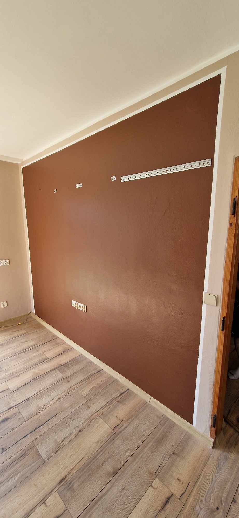 Czekoladowa ściana z widocznymi gniazdkami i listwą montażową, świeżo po malowaniu, z drewnopodobną podłogą i białym wykończeniem sufitu.