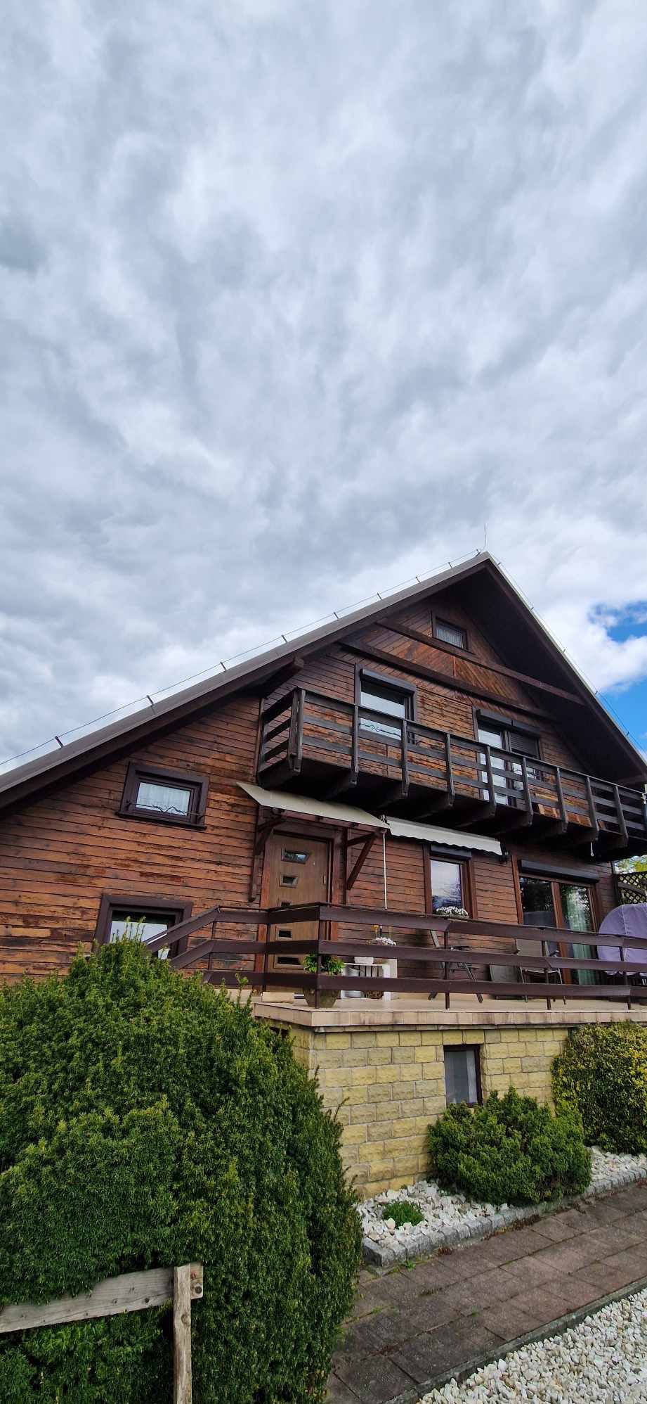 Dwupiętrowy dom z bali z dwoma balkonami, widok z dołu na tle pochmurnego nieba.