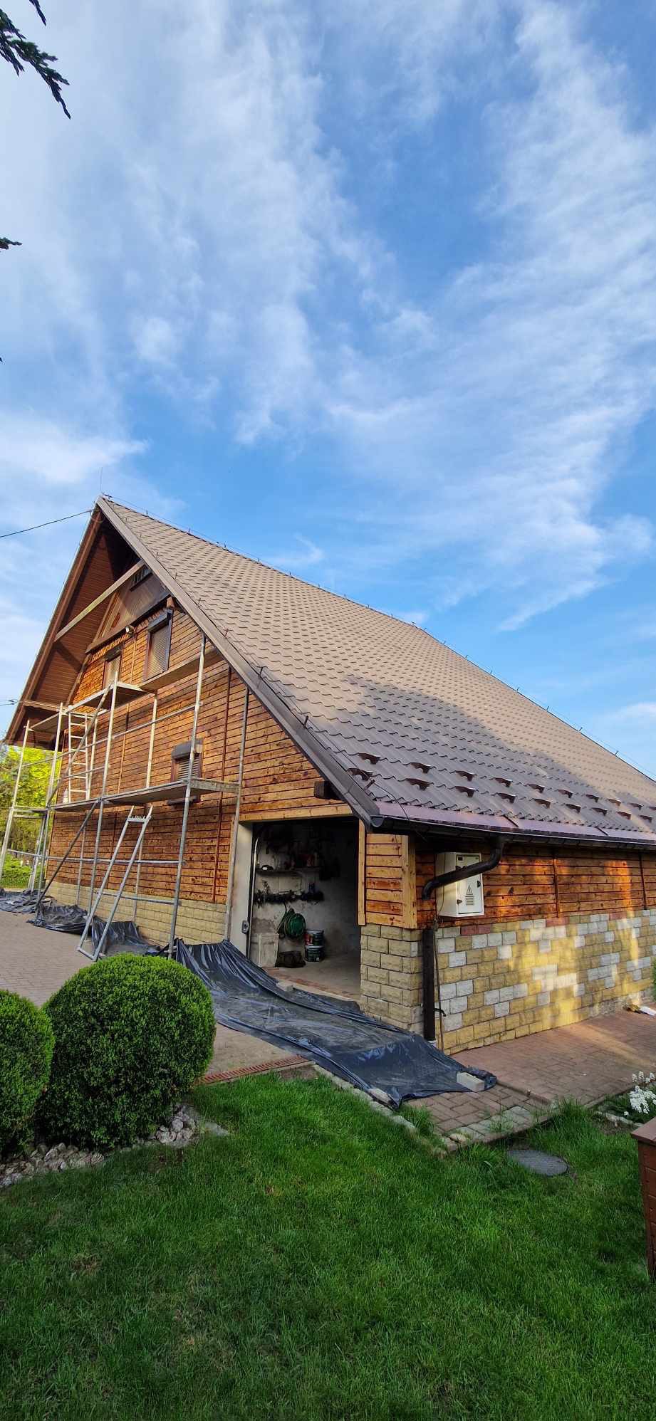 Drewniany budynek z nowym brązowym dachem, częściowo pokryty rusztowaniem, z widoczną elewacją z cegły i otwartym garażem z narzędziami wewnątrz, zabezpieczony folią ochronną.