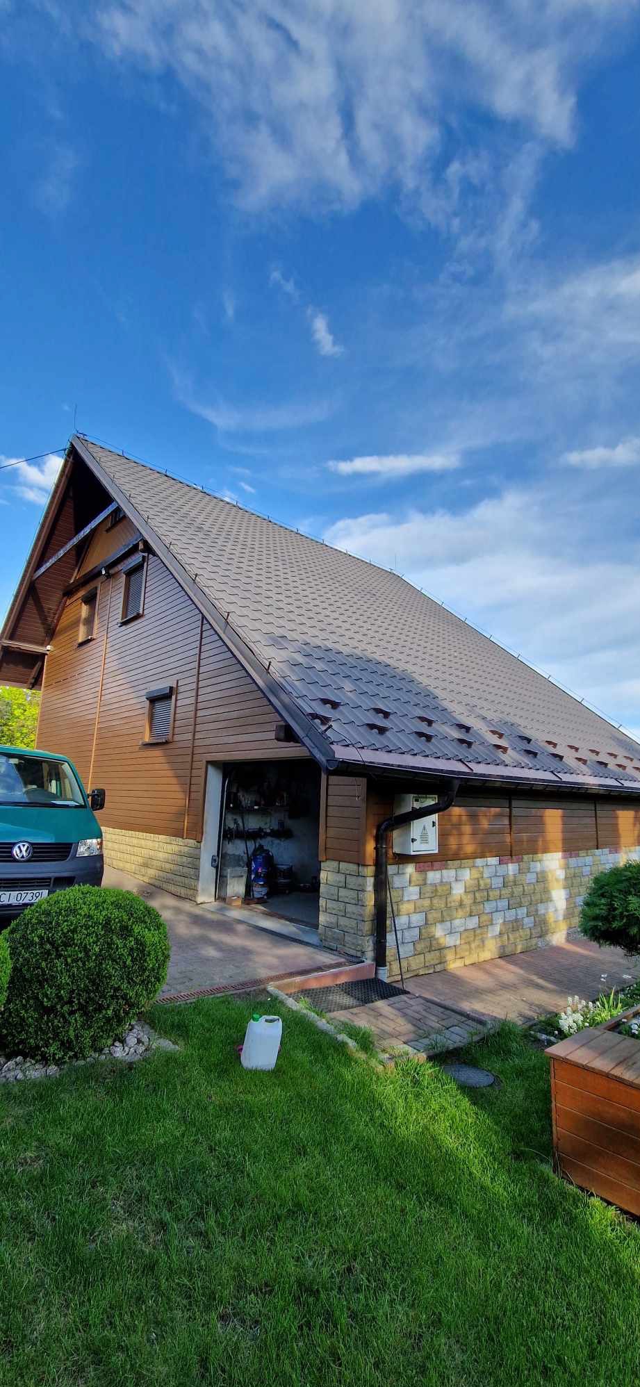 Dom jednorodzinny z brązową elewacją, grafitowym dachem i otwartym garażem, widziany z poziomu trawnika z krzewami i zielonym vanem z przodu.