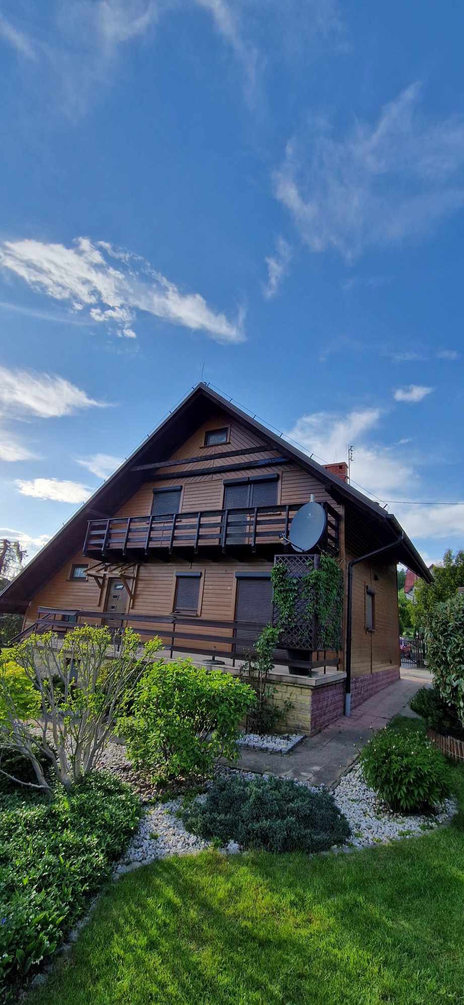 Drewniany dom z balkonem, brązową elewacją i satelitą na ścianie, otoczony zielenią w słoneczny dzień.