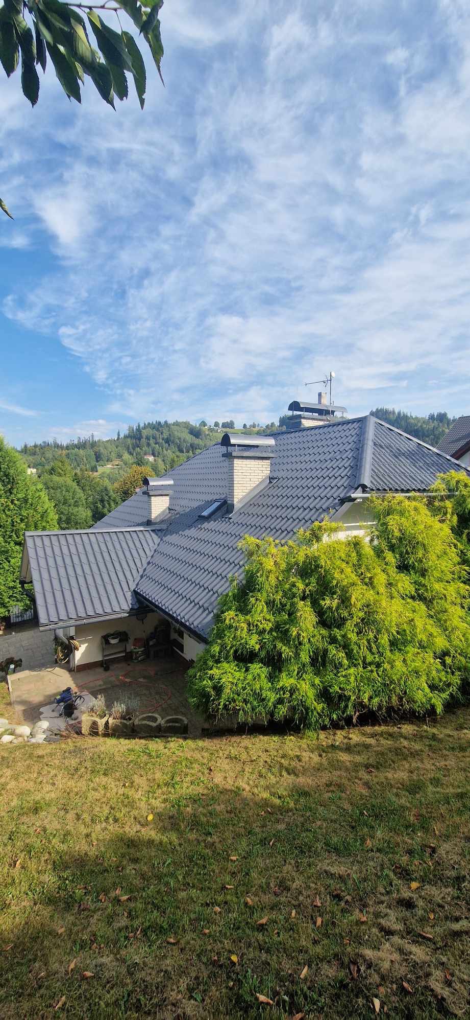 Widok z góry na dom z ciemnoszarą dachówką, ceglanymi kominami i bujną zielenią w tle, z widocznymi pagórkami i błękitnym niebem z chmurami.