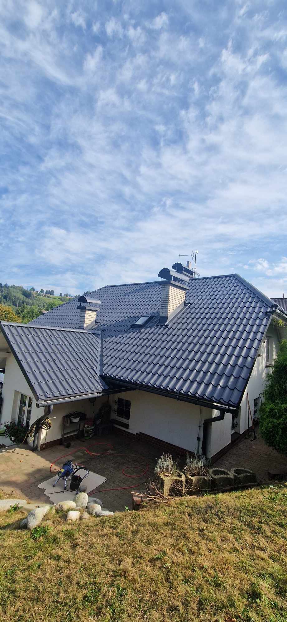 Dom jednorodzinny z ciemną dachówką ceramiczną, widok z góry, na podwórku leży rozłożony sprzęt do malowania natryskowego.