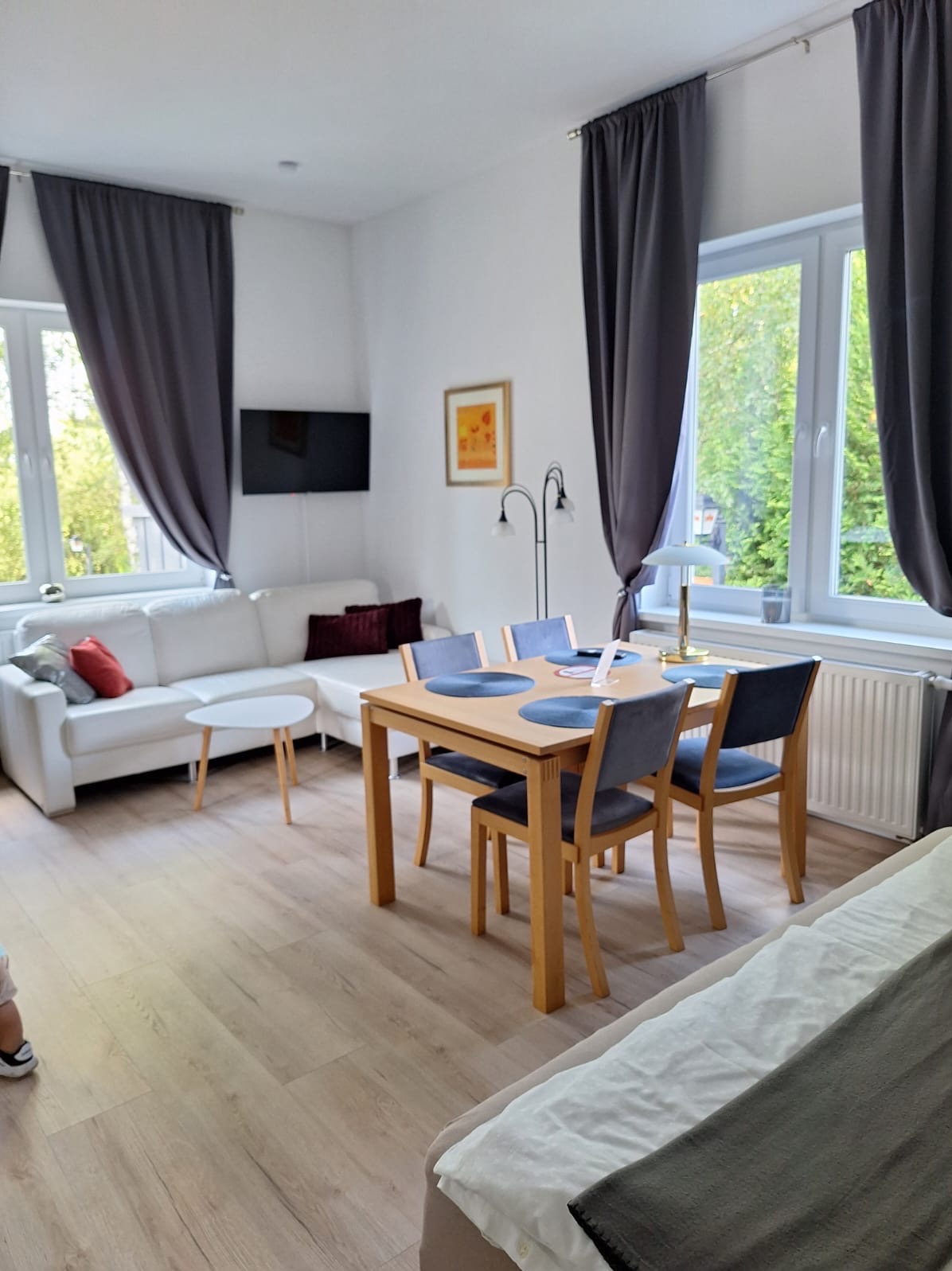 Jasne wnętrze pokoju dziennego z drewnianą podłogą, białym narożnikiem, stołem z szarymi krzesłami i szarymi zasłonami w oknach. Na ścianie wisi telewizor i obrazek.