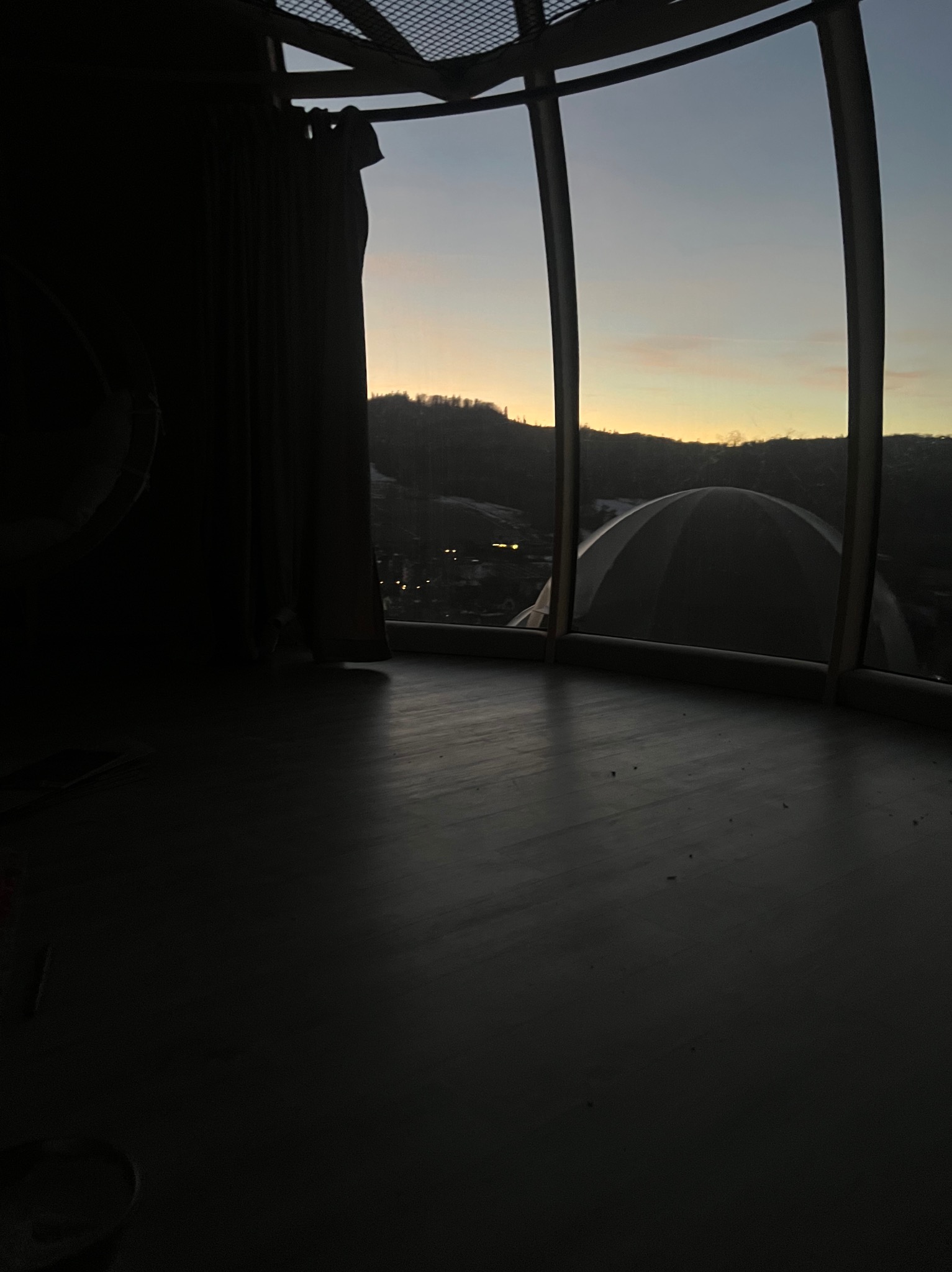 Wnętrze okrągłego pomieszczenia z dużymi oknami, widok na ośnieżone wzgórza i zachodzące słońce, drewniana podłoga w ciemnym odcieniu.