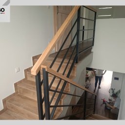 WBO Oskar Winowiecki - Nowoczesne schody z drewnianymi stopniami i czarną, metalową konstrukcją, widoczne z góry, z osobą pracującą na dole. Minimalistyczny design.