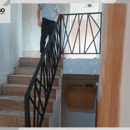 WBO Oskar Winowiecki - Nowoczesne, czarne poręcze schodowe z geometrycznym wzorem, zamontowane w jasnym wnętrzu. Widoczne drewnopodobne stopnie i fragment osoby na schodach.