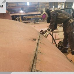 WBO Oskar Winowiecki - Spawacz w kombinezonie i masce spawalniczej łączy metalowe elementy konstrukcji w hali produkcyjnej. Widoczny sprzęt spawalniczy i fragment spawanej powierzchni.