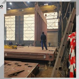 WBO Oskar Winowiecki - Wnętrze hali produkcyjnej z konstrukcją stalową w trakcie montażu. Widoczni pracownicy i drabina. Oświetlenie przemysłowe, surowy wygląd.