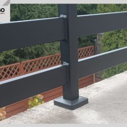 WBO Oskar Winowiecki - Fragment czarnej, metalowej konstrukcji z profili kwadratowych, prawdopodobnie balustrady, z widocznymi spawami i mocowaniem do podłoża. W tle zieleń i drewniane ogrodzenie.