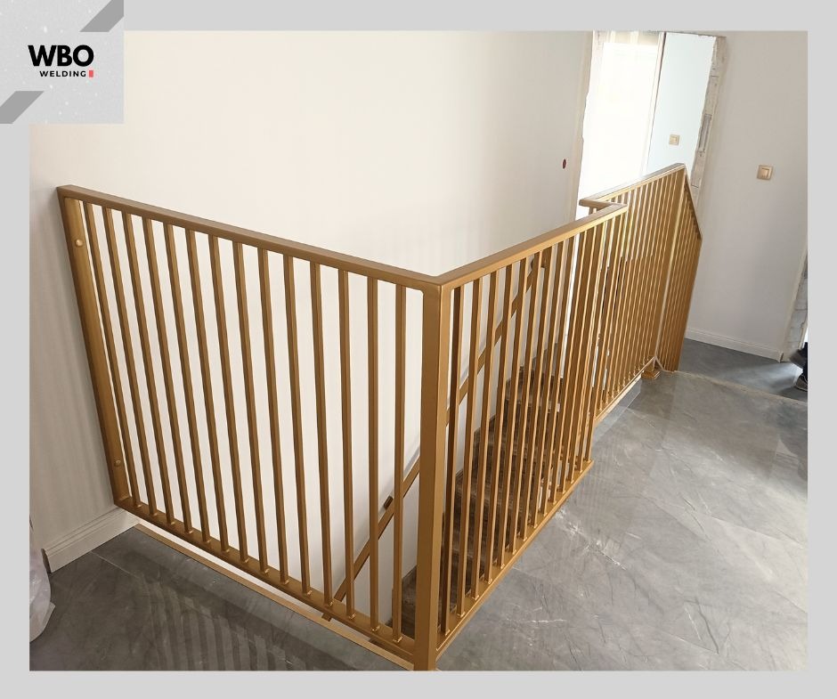 Złota, metalowa balustrada schodowa z pionowymi szczeblami, zamontowana w jasnym wnętrzu. Widok z góry na marmurowe schody. Minimalistyczny design.