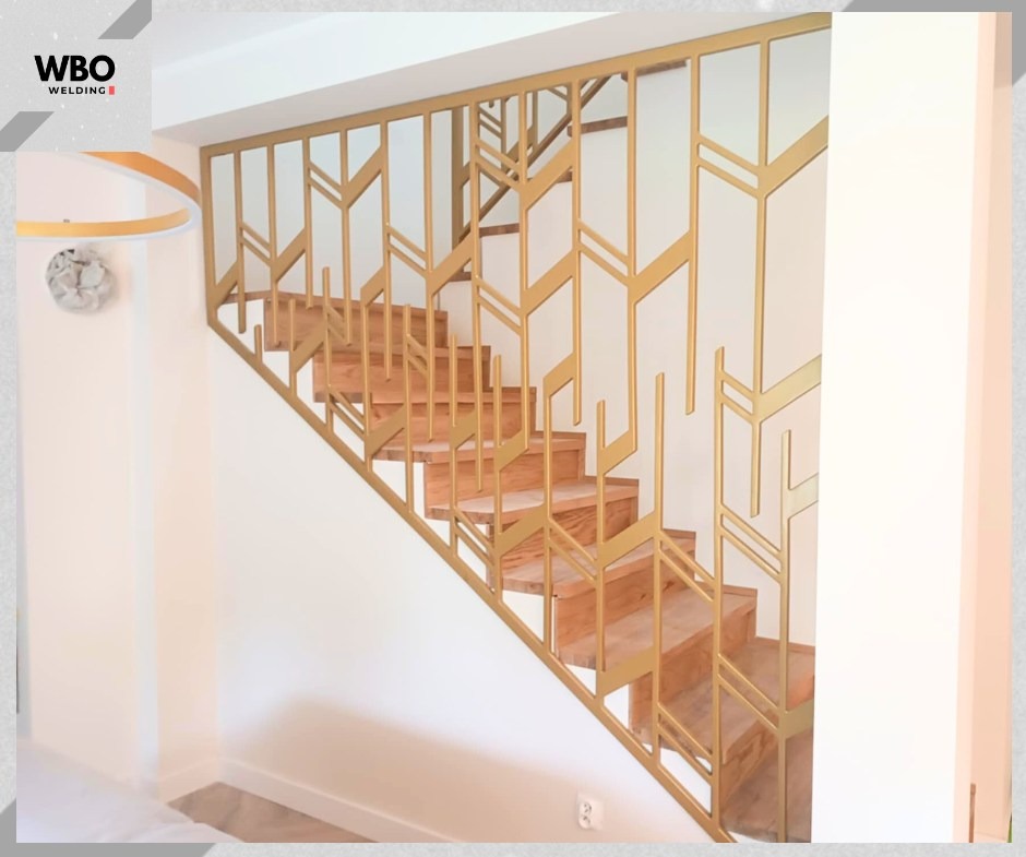 Złota, metalowa konstrukcja schodowa z geometrycznym wzorem, widoczna z boku, oświetlona naturalnym światłem. Schody drewniane, jasne ściany. Minimalistyczny design.