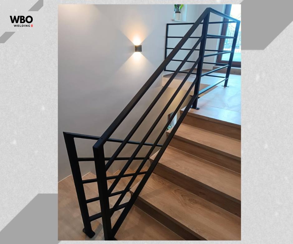 Nowoczesne, czarne poręcze schodowe z metalu, z ukośnymi elementami dekoracyjnymi. Widoczne drewniane stopnie i minimalistyczne oświetlenie ścienne.