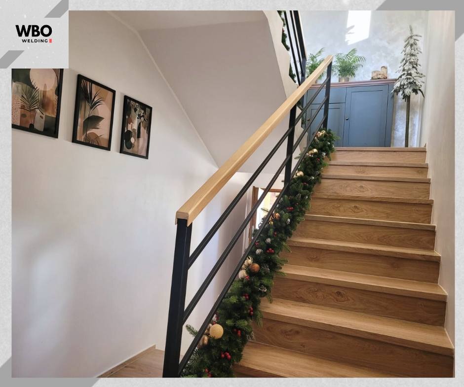 Schody drewniane z czarną metalową balustradą i jasnym drewnianym poręczą, udekorowane świąteczną girlandą. W tle biała ściana z obrazami i niebieska komoda.