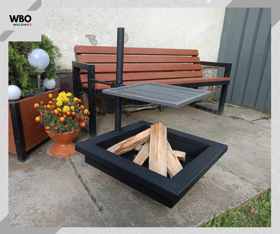 Metalowe palenisko z rusztem do grillowania, ustawione na betonowym podłożu, z drewnem w środku. W tle ławka i dekoracje ogrodowe.