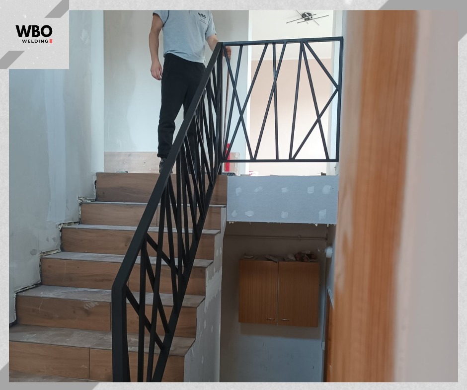 Nowoczesne, czarne poręcze schodowe z geometrycznym wzorem, zamontowane w jasnym wnętrzu. Widoczne drewnopodobne stopnie i fragment osoby na schodach.