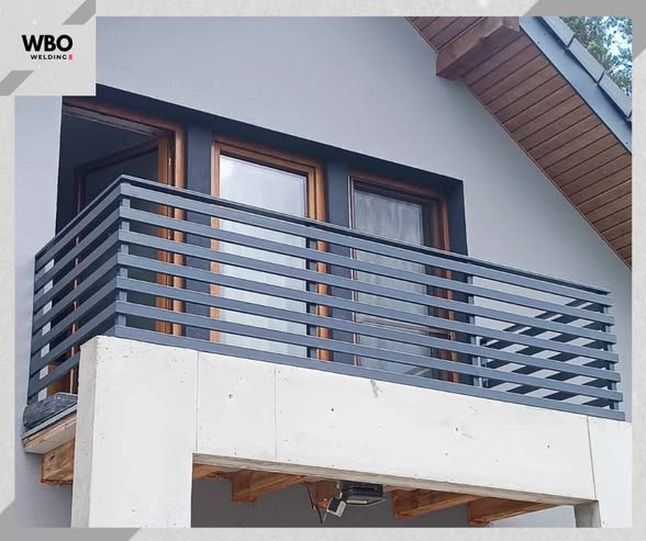 Nowoczesna, pozioma, szara konstrukcja zabezpieczająca balkon, z widocznymi oknami i fragmentem elewacji domu w tle. Minimalistyczny design.