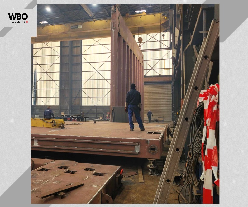 Wnętrze hali produkcyjnej z konstrukcją stalową w trakcie montażu. Widoczni pracownicy i drabina. Oświetlenie przemysłowe, surowy wygląd.