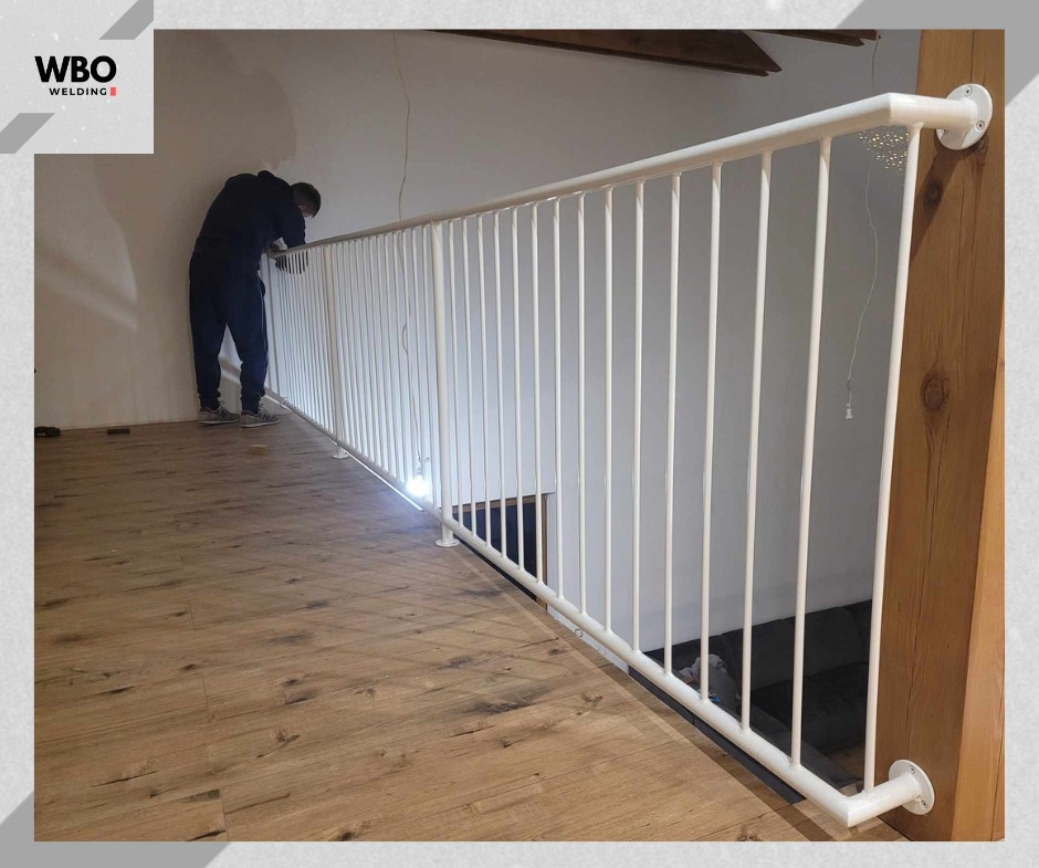 Biała, metalowa barierka na antresoli z drewnianą podłogą. Osoba w ciemnym ubraniu pochyla się nad nią. Widoczny fragment drewnianej belki konstrukcyjnej.