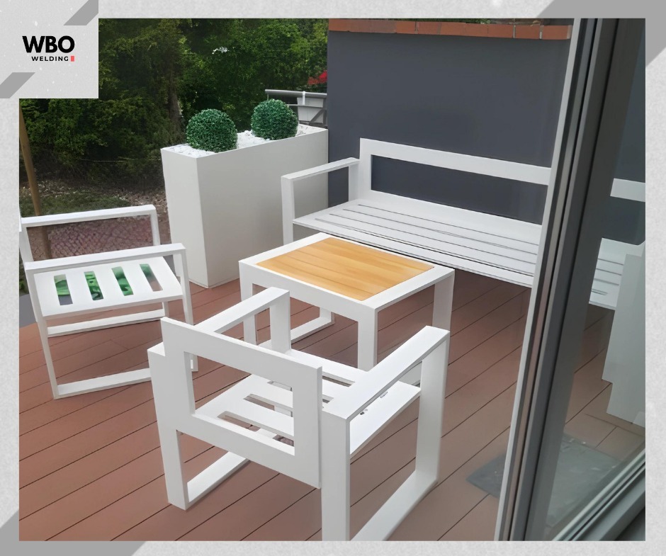 Białe, metalowe meble ogrodowe (krzesła, ławka, stolik z drewnianym blatem) na tarasie z desek kompozytowych. Minimalistyczny design, nowoczesny styl.
