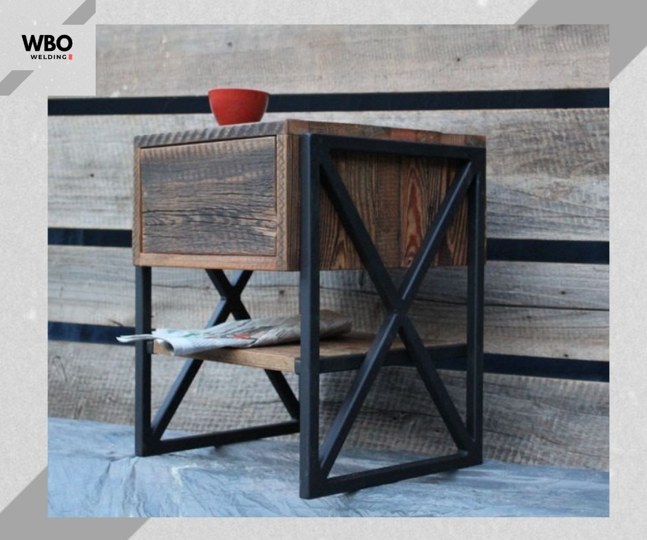 Stolik nocny z drewna i metalu w stylu industrialnym, wykonany w Bydgoszczy. Widoczna czerwona miseczka na blacie i gazeta na półce. Metalowa konstrukcja w kształcie litery X.