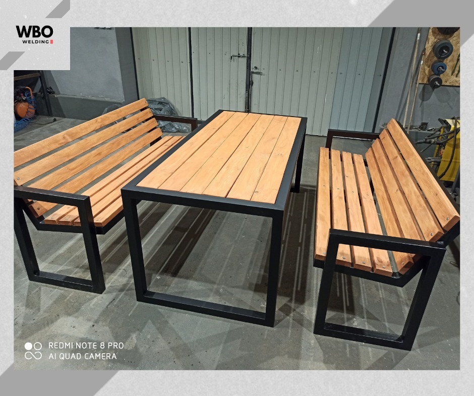 Komplet mebli ogrodowych: stół i dwie ławki z drewnianymi siedziskami i blatami, osadzonych na czarnych, metalowych ramach o surowym, industrialnym wyglądzie.