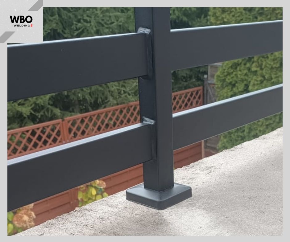 Fragment czarnej, metalowej konstrukcji z profili kwadratowych, prawdopodobnie balustrady, z widocznymi spawami i mocowaniem do podłoża. W tle zieleń i drewniane ogrodzenie.