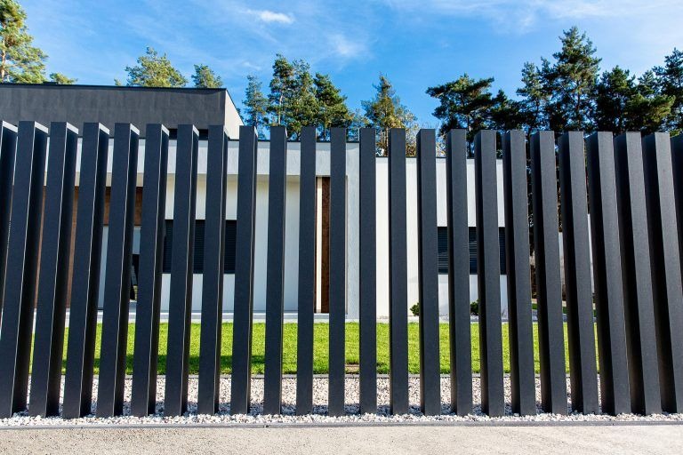 Nowoczesne, pionowe ogrodzenie z ciemnych, metalowych słupków na tle domu z ogrodem. Minimalistyczny design, równomierne odstępy, żwirowe podłoże.