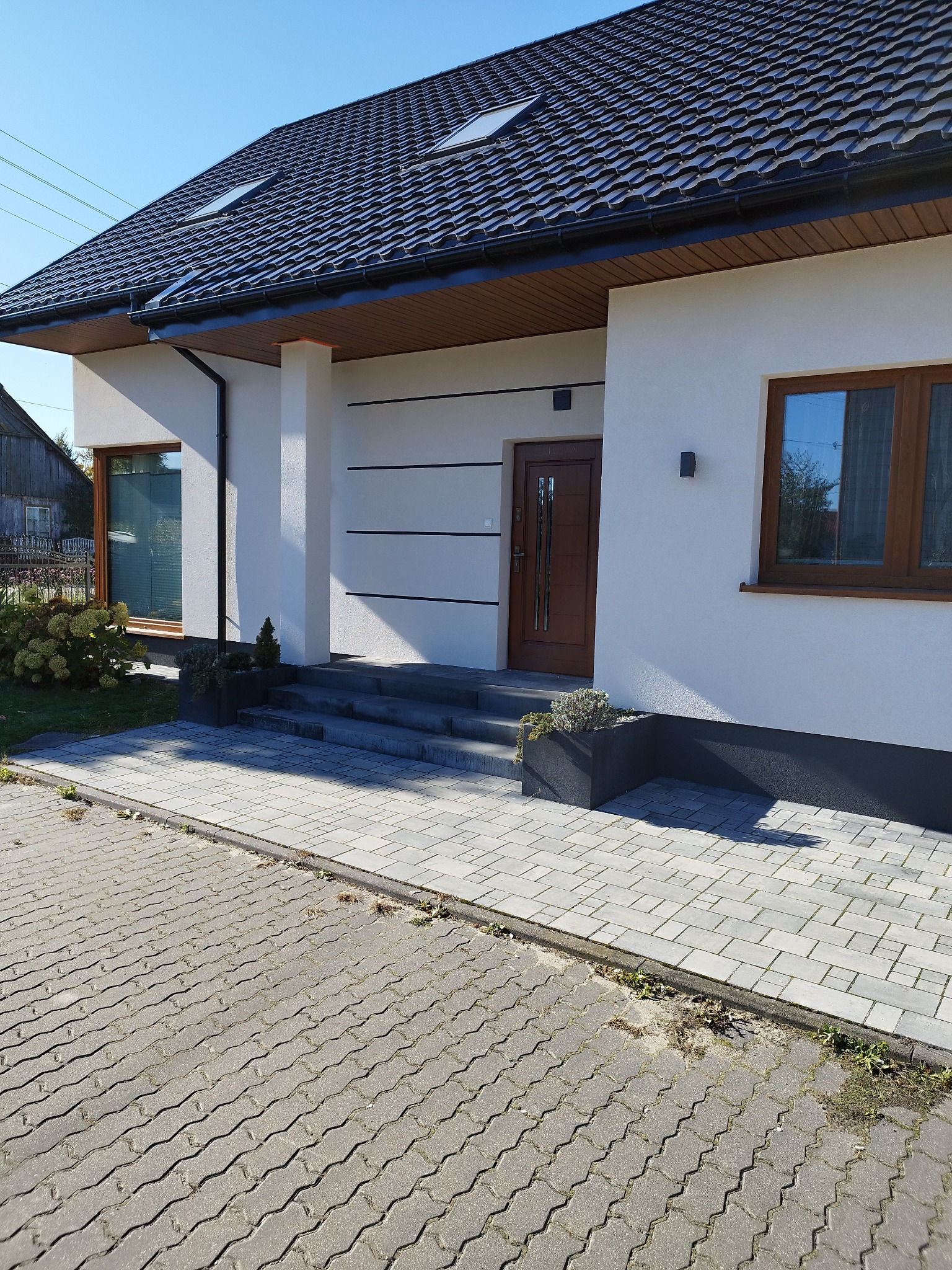 Nowoczesny dom jednorodzinny z białą elewacją, brązowymi oknami i drzwiami wejściowymi, oraz ciemną dachówką. Widoczne trzy poziome pasy dekoracyjne na elewacji. Ułożona kostka brukowa przed wejściem.