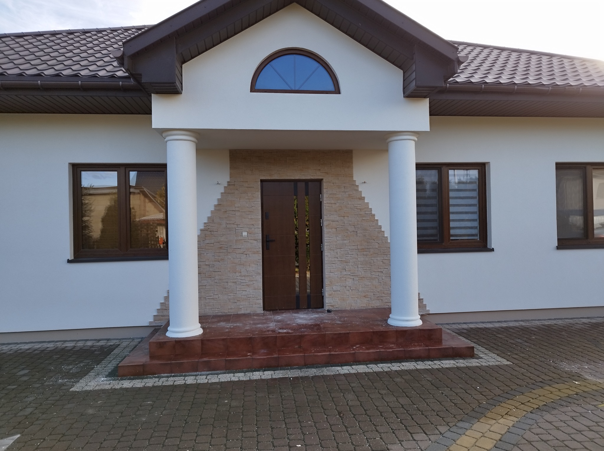 Wykończone wejście do domu z białymi ścianami, kolumnami i kamienną okładziną wokół brązowych drzwi wejściowych, widoczne brązowe okna i dachówka.