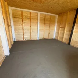 Świeżo wylana, mokra posadzka betonowa wewnątrz drewnianej konstrukcji budynku, widoczne drewniane ściany i sufit z płyty OSB, białe słupy konstrukcyjne.