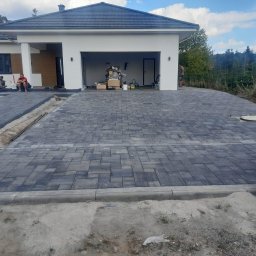 Prestivo Cezary Mokrzycki - Nowo ułożona, szara kostka brukowa przed nowoczesnym domem z garażem; widoczne materiały budowlane i poziomica.