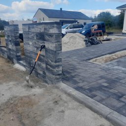 Prestivo Cezary Mokrzycki - Fragment posesji w trakcie prac: wznoszony mur z szarej, ciosanej kostki betonowej, ułożona szara kostka brukowa, widoczna łopata oparta o mur, w tle domy i samochody.