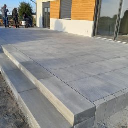 Prestivo Cezary Mokrzycki - Nowoczesny taras z szarych płyt betonowych z dwustopniowymi schodami, ekipa budowlana w tle, widok na elewację domu z drewnianymi elementami.