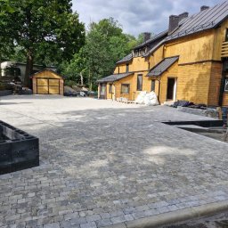 Prestivo Cezary Mokrzycki - Nowo wyłożony dziedziniec z szarej kostki brukowej przed drewnianym domem z ciemnym dachem; widoczny miniładowarka, ogrodzenie z betonowych bloczków i worki z materiałami budowlanymi.