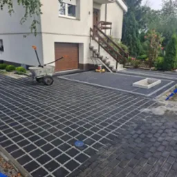 Ułożona nawierzchnia z ażurowych, czarnych płyt betonowych z wypełnieniem żwirowym, widoczny wózek budowlany, zagęszczarka wibracyjna, fragment domu z garażem i schodami wejściowymi, oraz rząd tuj.