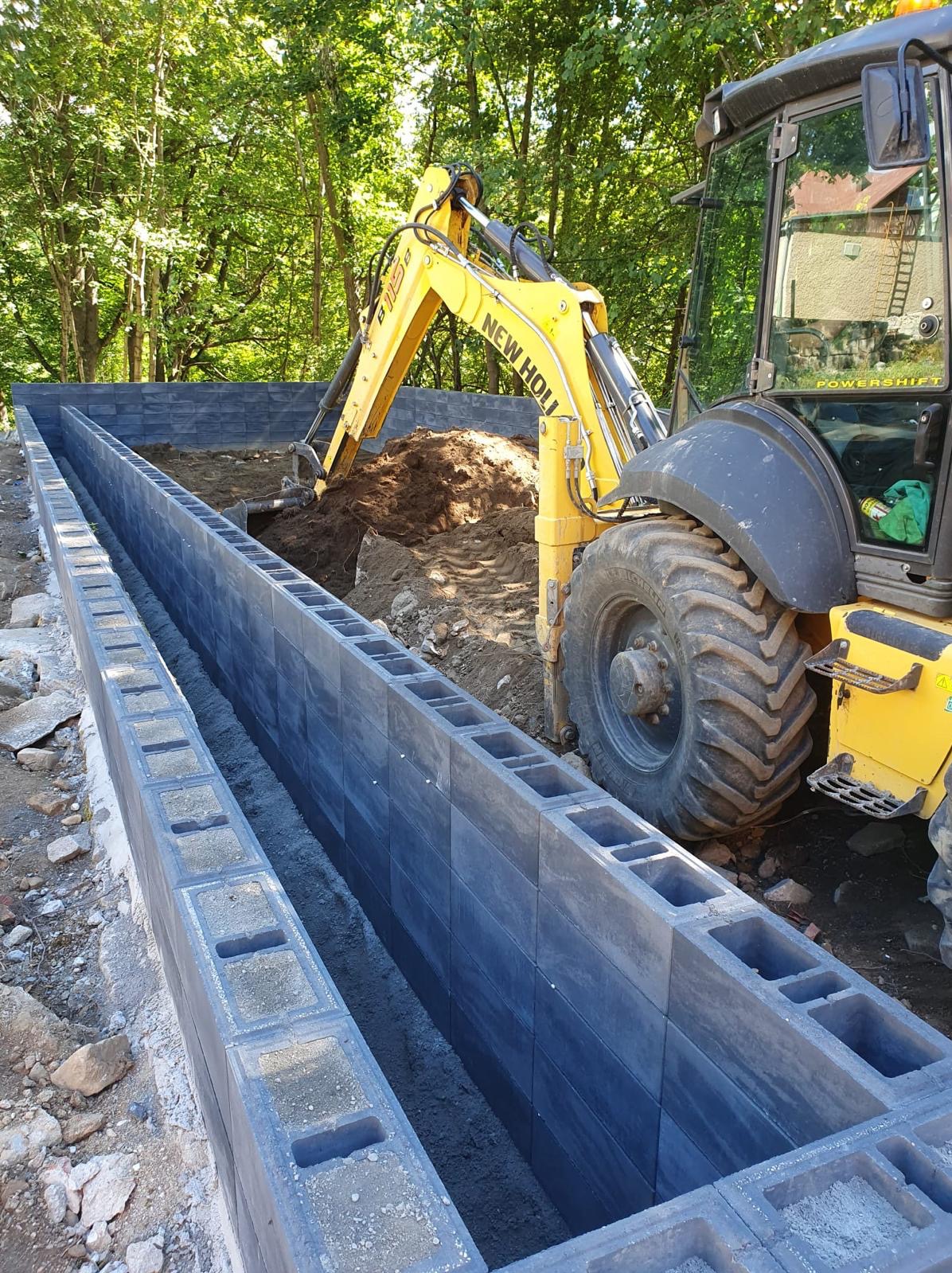 Żółta koparko-ładowarka New Holland nabiera ziemię w wykopie wyłożonym szarymi bloczkami betonowymi z otworami, przygotowywanym pod fundament. Widoczne drzewa w tle.