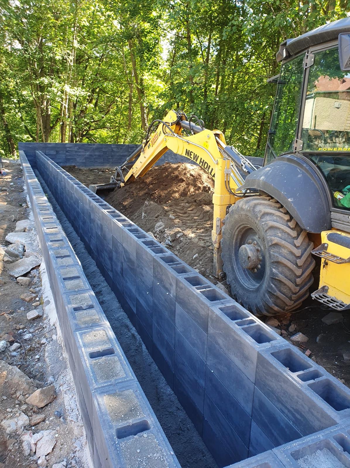 Żółta koparka New Holland zasypuje ziemią fundament z betonowych bloczków w lesistym otoczeniu.