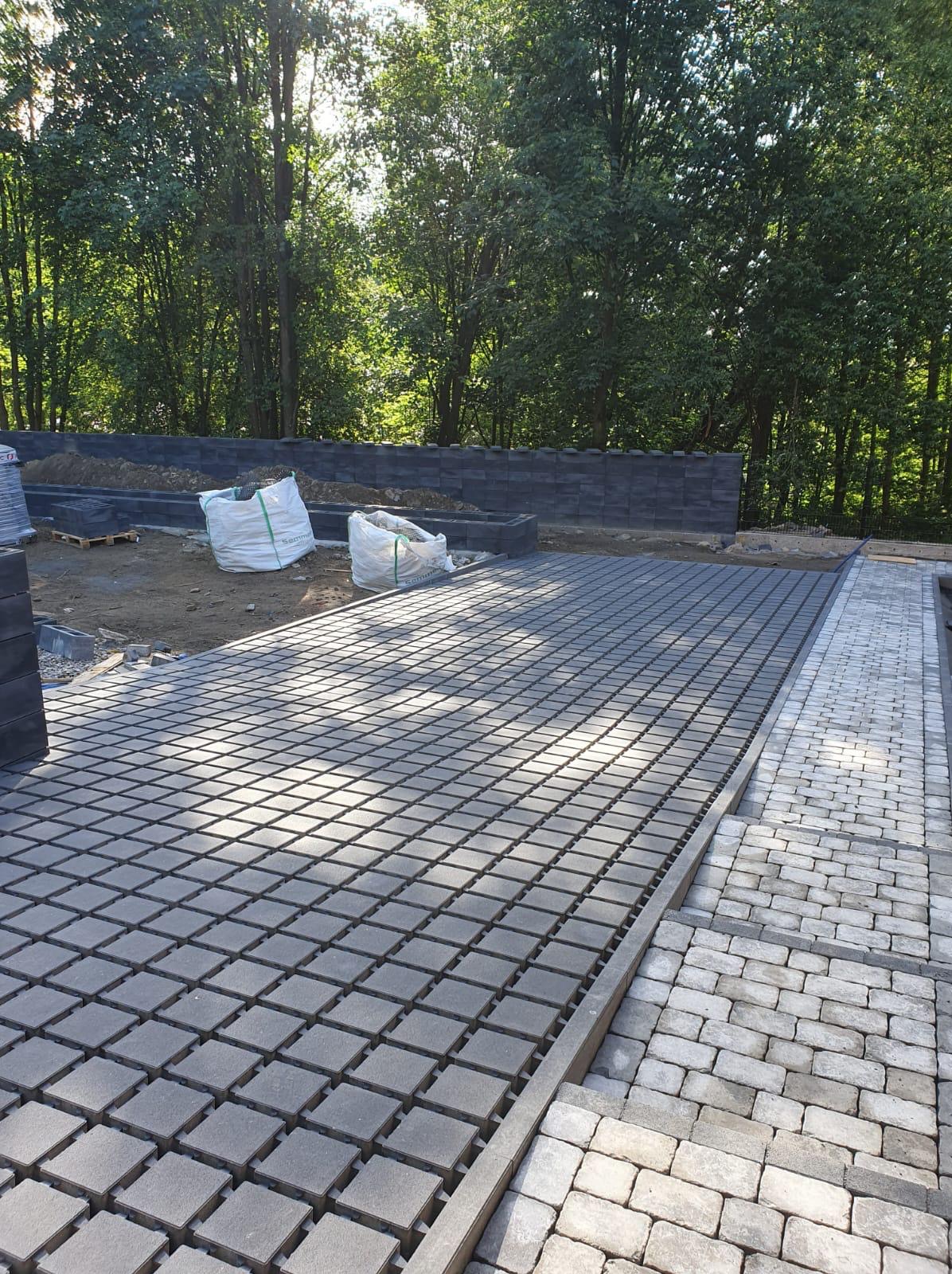 Ułożona nawierzchnia z szarej kostki brukowej kwadratowej z widoczną siatką dystansową, obok fragment chodnika z jasnej kostki prostokątnej, w tle worki z materiałem budowlanym i fragment murku.