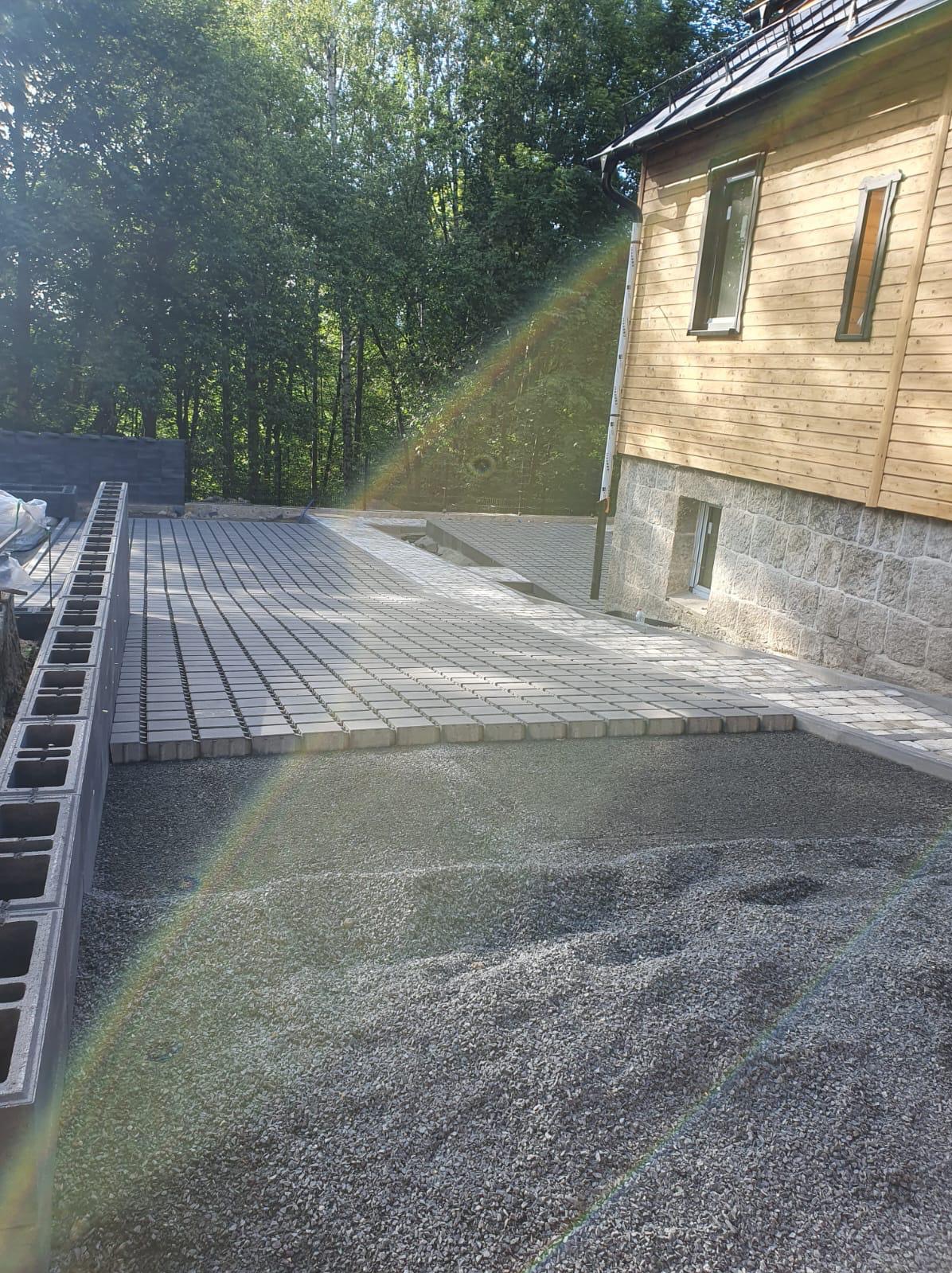 Fragment podjazdu w trakcie budowy z częściowo ułożoną kostką brukową, podsypką żwirową i betonowymi elementami obrzeża, dom z drewnianą elewacją w tle.
