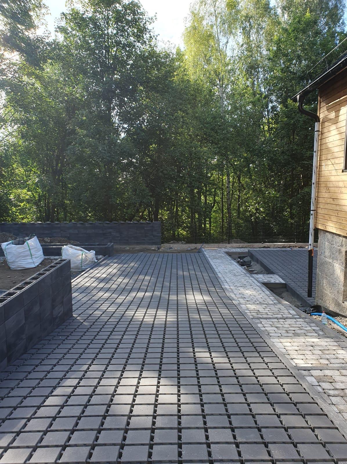 Układanie szarej kostki brukowej z wąskimi szczelinami na podjeździe, widoczne worki z materiałem budowlanym i fragment budynku z drewnianą elewacją w tle.