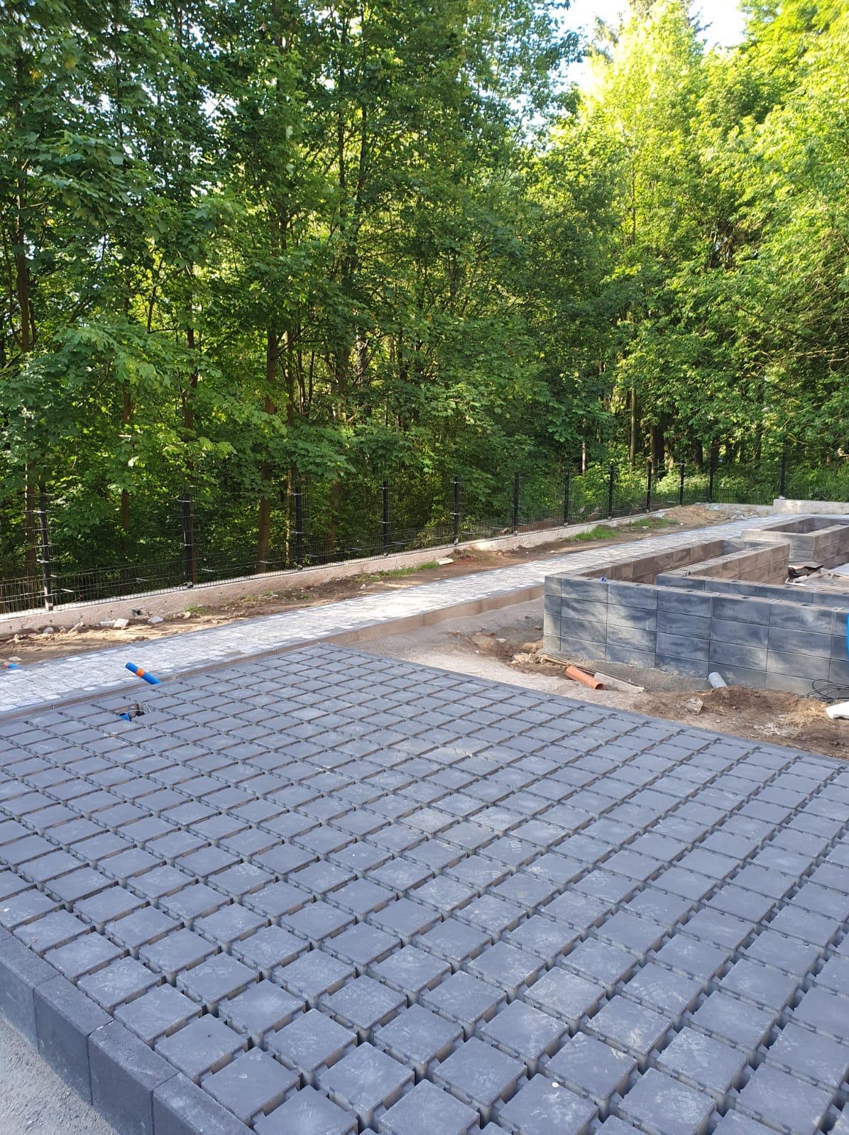 Szara kostka brukowa ułożona na placu budowy, w tle widoczny fragment ogrodzenia i drzewa, widać postęp prac.