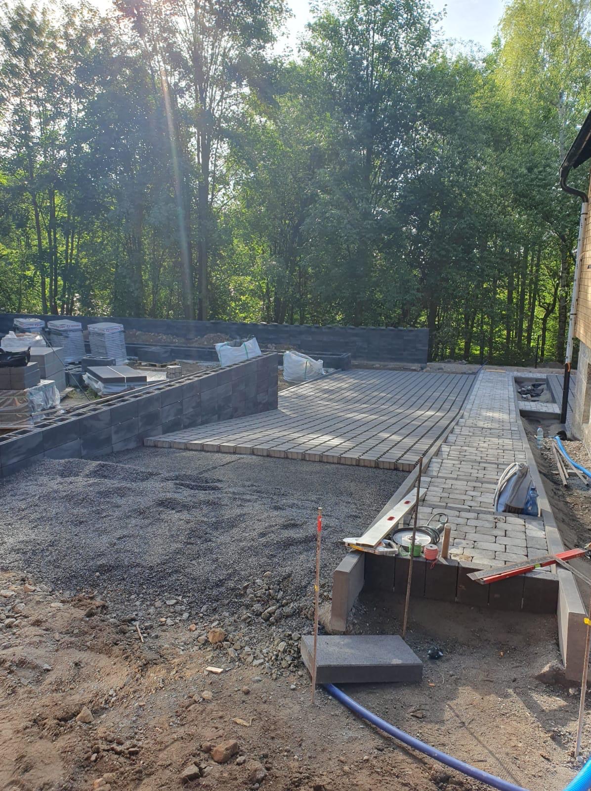 Fragment posesji w trakcie budowy tarasu i ścieżki z kostki brukowej, widoczne narzędzia, podsypka i palety z kostką w tle, drzewa