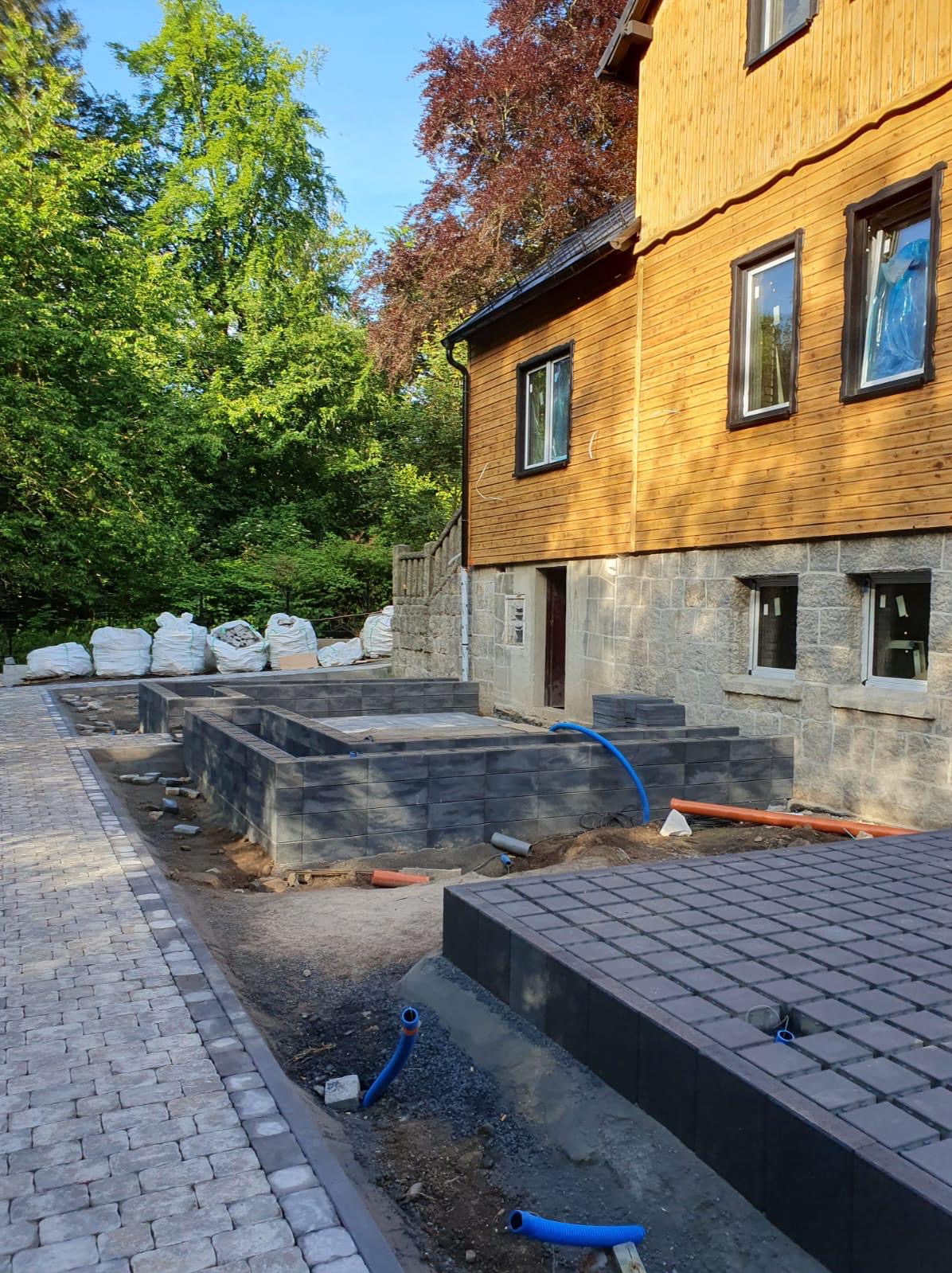 Budowa tarasu z kostki brukowej przy drewniano-kamiennym domu, widoczne fundamenty, worki z materiałem budowlanym i fragment istniejącego chodnika z kostki.