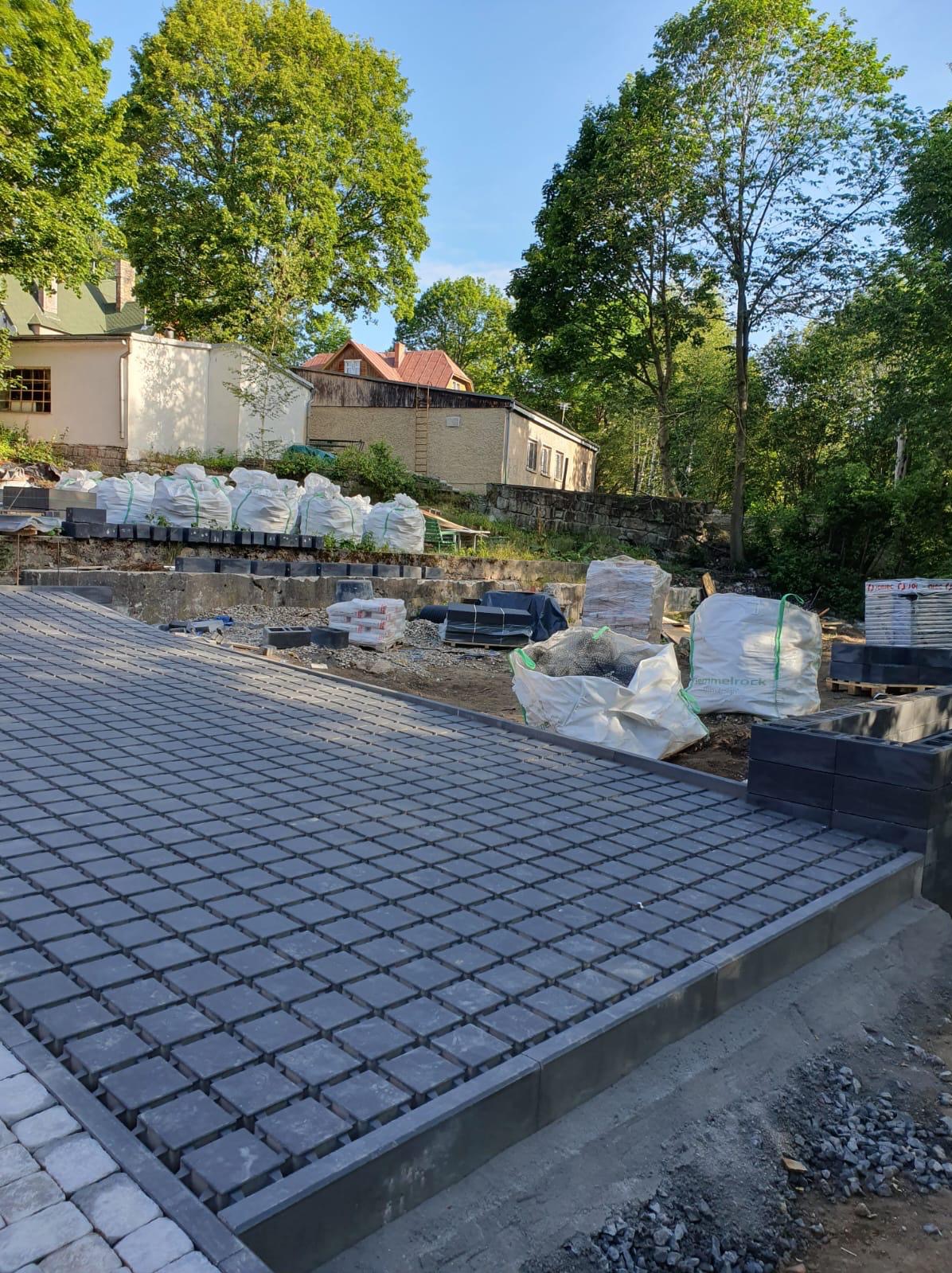 Częściowo ułożona, szara kostka brukowa na przygotowanym podłożu. W tle worki z materiałami budowlanymi, teren budowy i zieleń drzew.