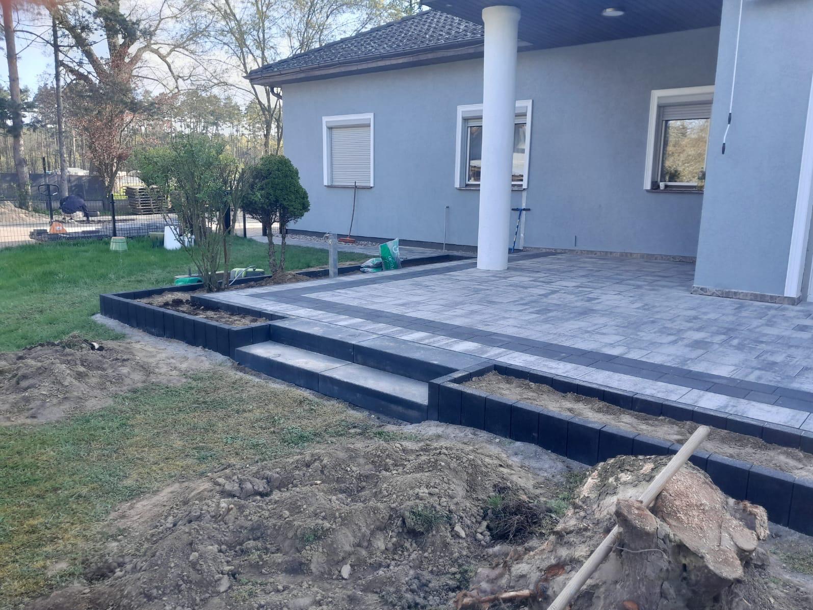 Częściowo ułożona szara kostka brukowa z czarnym obramowaniem, tworząca taras przed domem z kolumną. Widoczne schody z kostki i przygotowane rabaty ziemne, z pryzmą ziemi na pierwszym planie.