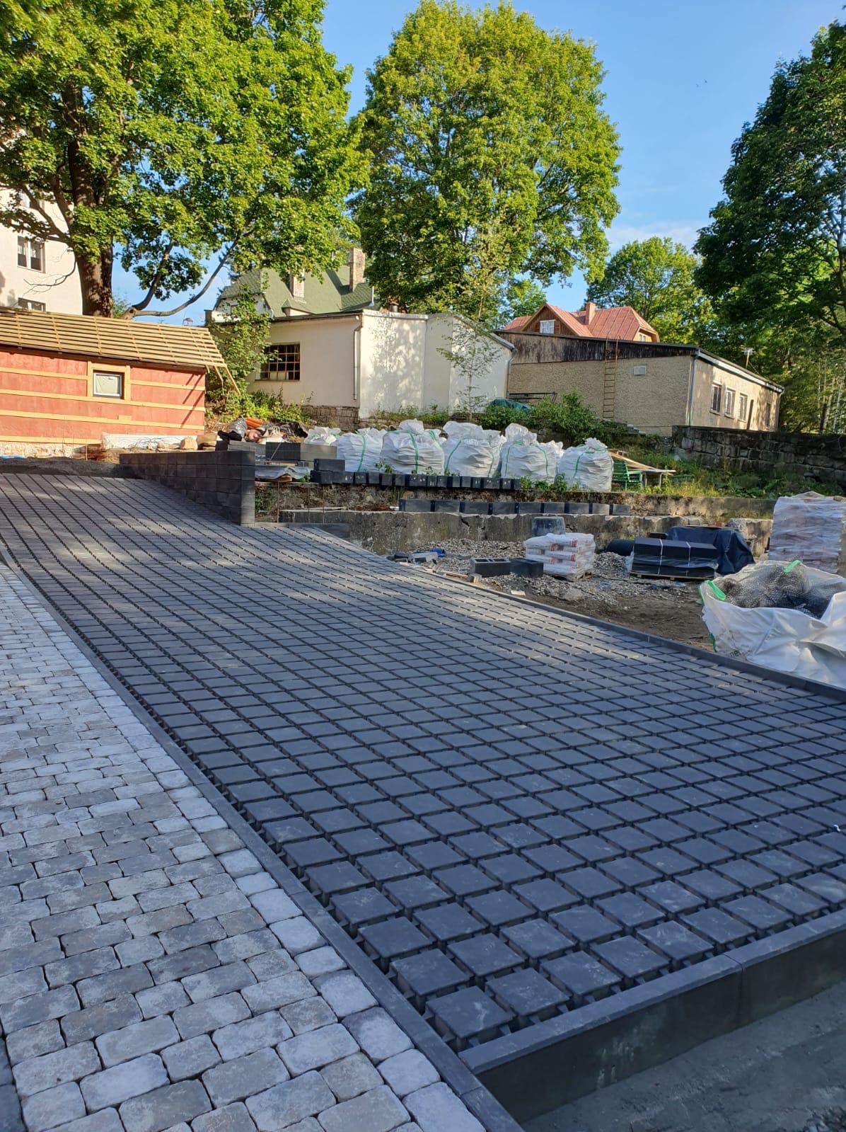 Ukończona podjazd z ciemnej kostki brukowej na posesji, z widocznymi workami materiałów budowlanych w tle i budynkami mieszkalnymi.