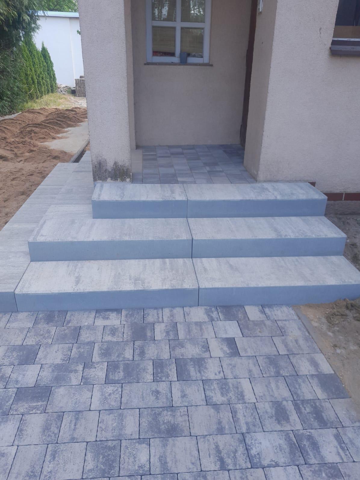 Nowo ułożone, szare płytki brukowe prowadzące do wejścia domu z trzema stopniami wykonanymi z betonowych bloków.